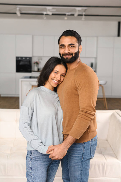 Smiling man hugging his charming woman. Two people standing and joyfully looking at the camera. Young Indian couple happily spending time in cozy kitchen - Foto, imagen