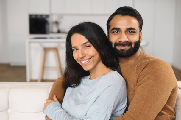 Close-up of happy interracial couple posing while sitting at the sofa with kitchen background. Happy owners of new flat embracing in front of the camera - Foto, imagen