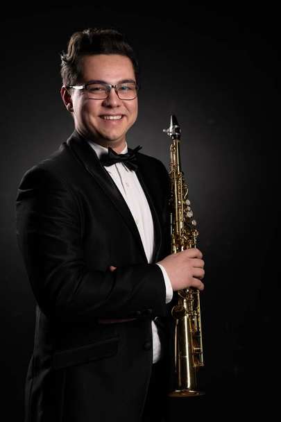 Japanese Musician Saxophonist in a Black Classic Suit Holds a Soprano Straight Saxophone in his Hand and Poses for the Camera on a Black Background. Close-up Portrait  - Foto, Bild
