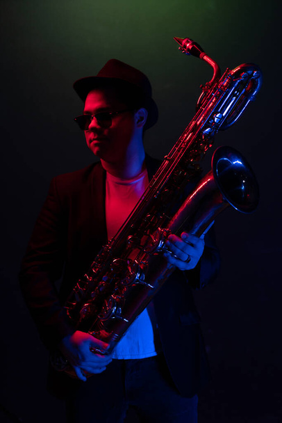 Young Saxophonist in a Black Jacket, Hat and Sunglasses Holds a Baritone Saxophone. Dark Background. Neon Light. Close-up Portrait - Foto, afbeelding