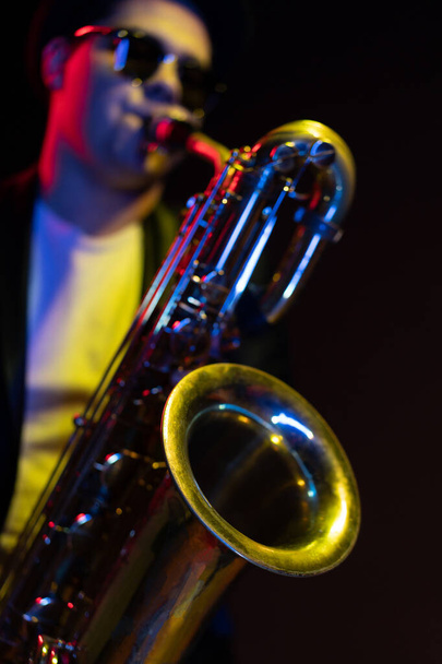 Saxophonist Guy in Sunglasses Plays the Tenor Saxophone, Musician Blows the Saxophone. Selective Focus on Bell of Saxophone. Blurred Background, Neon Light - Foto, imagen
