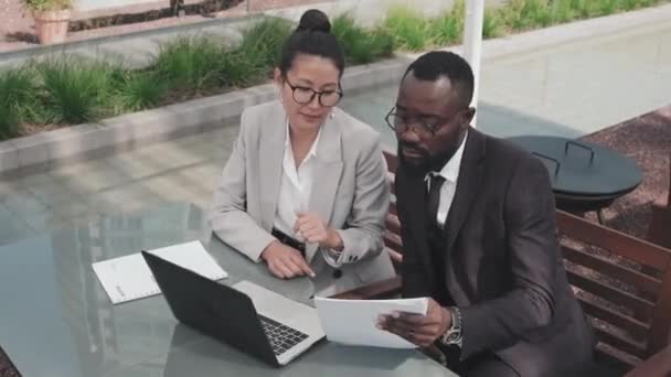 Tracking hoge hoek schot van Aziatische zakenvrouw en Afro-Amerikaanse zakenman in formele kleding zitten aan tafel in de stoep cafe in de stad en bespreken van het werk - Video