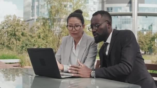 Śledzenie zdjęcia azjatyckiej bizneswoman i Afroamerykańskiego biznesmena w formalnym ubraniu siedzi na zewnątrz patio kawiarni i omawia pracę patrząc na laptopa - Materiał filmowy, wideo