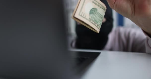 Киберпреступник в черной маске держит доллары на крючке и смотрит в ноутбук - Кадры, видео