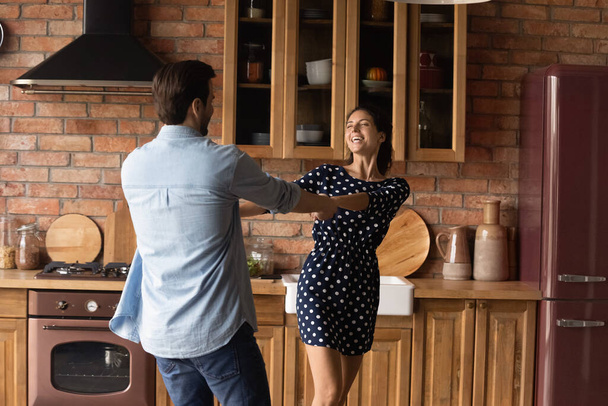 Joyful laughing millennial couple dancing together in kitchen. - Фото, изображение
