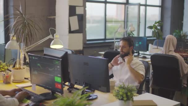 Nagy látószögű szakállas Biracial férfi ül az íróasztalnál az irodában, számítógép, üzembe over-ear fülhallgató, munkatársak beszél nappal, az emberek dolgoznak informatikai cég - Felvétel, videó