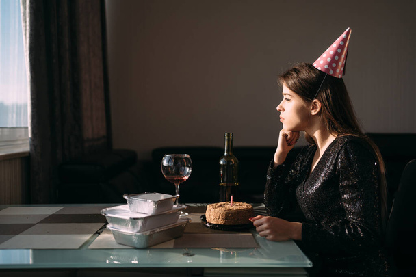 Young woman sitting alone at the table on her birthday. festive hat on the head. The lady feels bored and lonely. A bottle of wine and a birthday cake. Sad and lonely birthday concept. - Photo, Image
