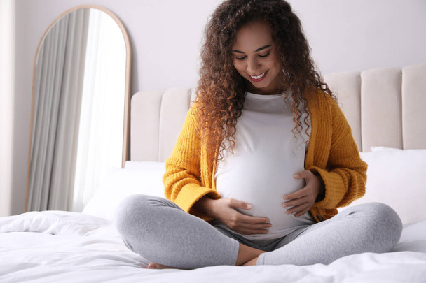 Pregnant young African-American woman sitting on bed at home - Foto, afbeelding