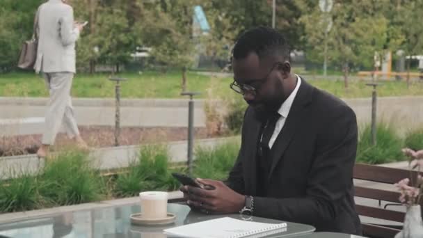 Handheld tracking shot of African-American businessman in suit and glasses sitting at table in sidewalk cafe and typing on mobile phone - Filmati, video