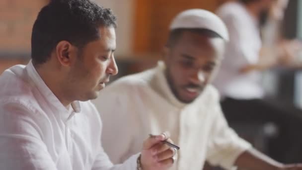 Medio primer plano del joven musulmán negro y su socio de negocios de Oriente Medio se reúnen en la cafetería, se sientan a la mesa, hablan y trabajan con documentos - Imágenes, Vídeo