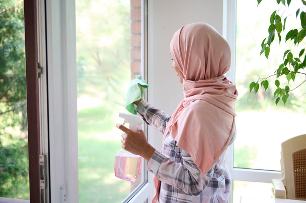 Mulher muçulmana árabe com a cabeça coberta por um hijab lava janelas, remove manchas, pulveriza detergente e limpa com um pano desfrutando de uma limpeza geral da primavera da casa com jardim no fundo - Foto, Imagem