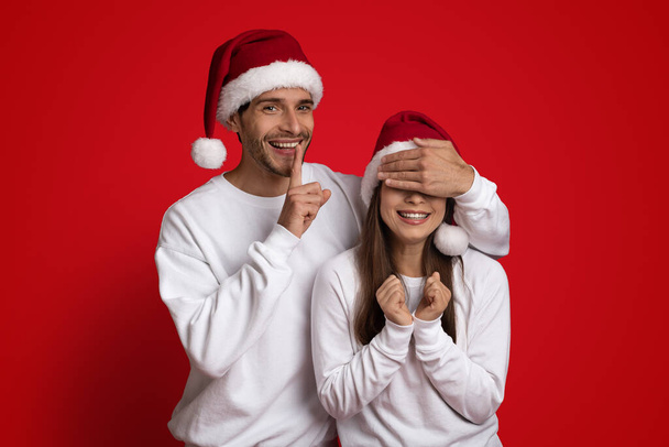 Весела людина в Санта капелюсі покриває подруги очі і показує знак
 - Фото, зображення