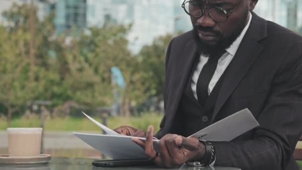 Handheld close up of busy African-American businessman in formal wear and glasses sitting at table in sidewalk cafe and looking through file with financial documents - Filmati, video