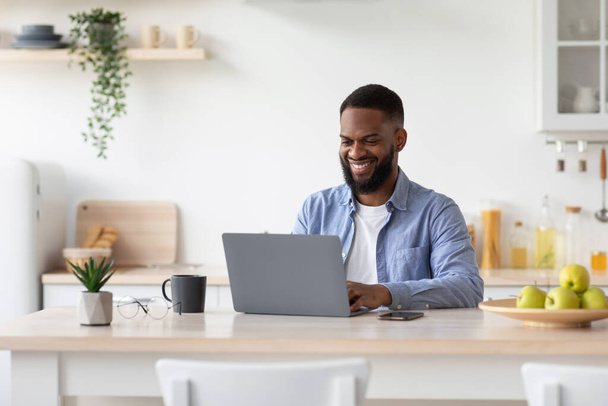 Feliz milenar afro-americano barbudo masculino trabalhando no laptop no interior da cozinha escandinava, espaço livre - Foto, Imagem