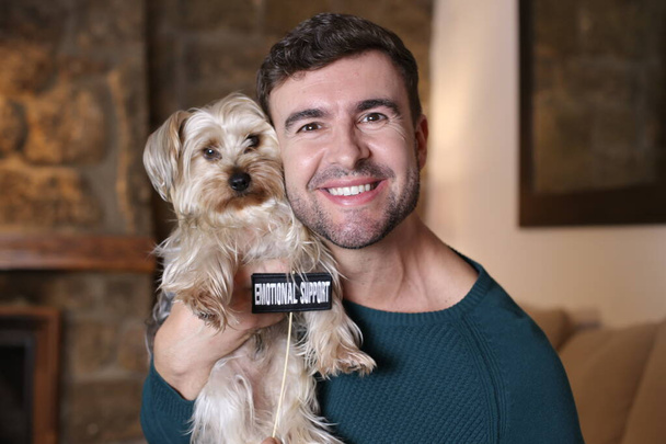 子犬を抱えているハンサムな若い男と家で感情的なサポートサインのクローズアップ肖像画 - 写真・画像