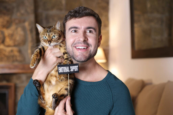 primer plano retrato de guapo joven sosteniendo su exótico gato y emocional apoyo signo en casa - Foto, imagen