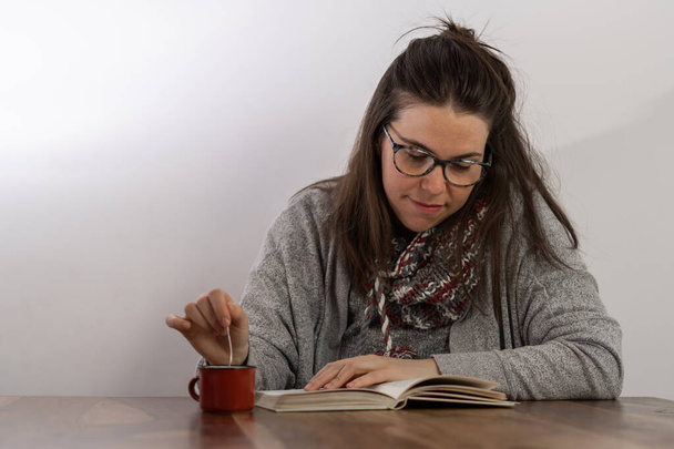 young brunette girl with long hair and glasses drinking a cup of coffee while reading a book seated at the table - Foto, Imagen