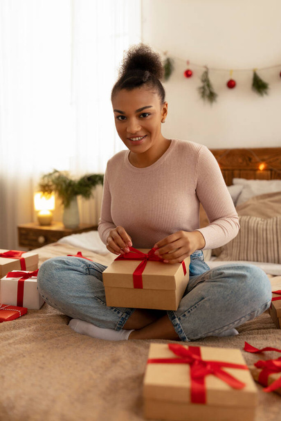 Handgemachte Geschenke. Glückliche schwarze Dame wickelt Geschenkboxen ein, macht Verbeugung mit rotem Bandseil, sitzt auf dem Bett - Foto, Bild