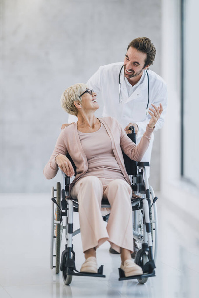 Shot of a young doctor pushing his senior wheelchair-bound woman patient and talking with her. - Fotografie, Obrázek