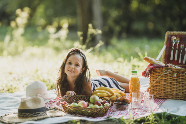 Una linda niña sonriente está tumbada en la manta sobre una hierba y disfrutando de un día de picnic en el parque.. - Foto, imagen