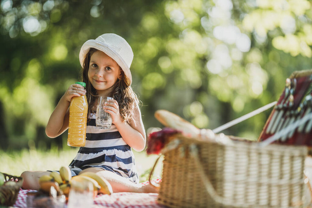 Une jolie petite fille souriante qui boit du jus d'orange tout en profitant d'une journée de pique-nique dans la nature. - Photo, image