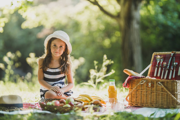 Cute uśmiechnięta dziewczynka siedzi na trawie i ciesząc się piknik dzień w parku. - Zdjęcie, obraz