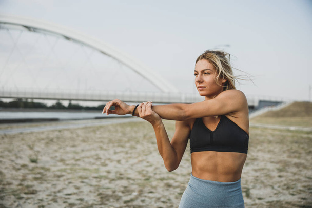 Spor yapan genç bir kadın şehirde nehre yakın bir yerde egzersiz yaparken ısınıyor ve kollarını geriyor.. - Fotoğraf, Görsel