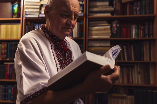 Uomo anziano in abiti popolari in possesso di un libro a casa biblioteca - Foto, immagini