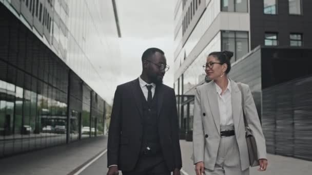 Tracking lövés vidám afro-amerikai üzletember és ázsiai üzletasszony a formális viselet sétál az utcán, és beszélgetni - Felvétel, videó