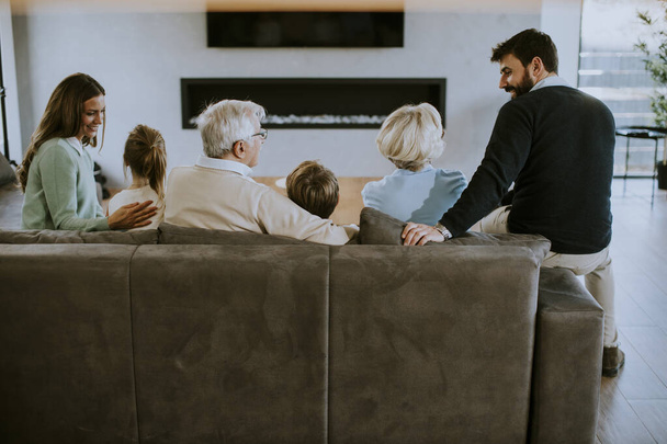 Семья из нескольких поколений сидит вместе на диване в современных апартаментах и смотрит телевизор - Фото, изображение
