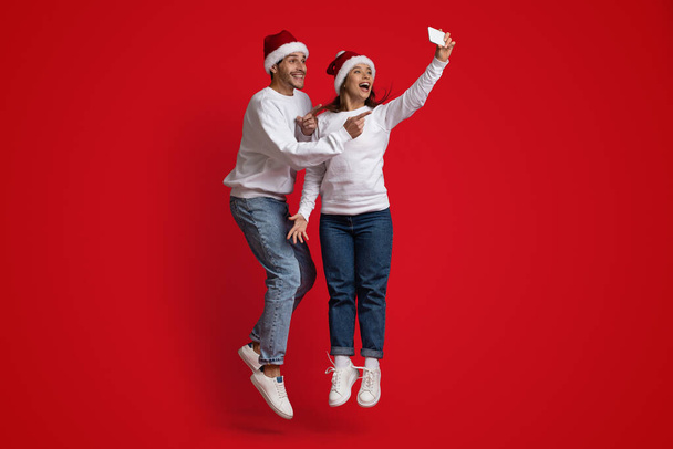 Noel Baba şapkası takan, zıplayan ve selfie çeken pozitifler. - Fotoğraf, Görsel