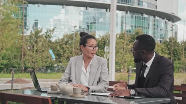 Medium shot of Asian businesswoman and African-American businessman signing documents while sitting at table in outdoor cafe, then shaking hands - Filmagem, Vídeo