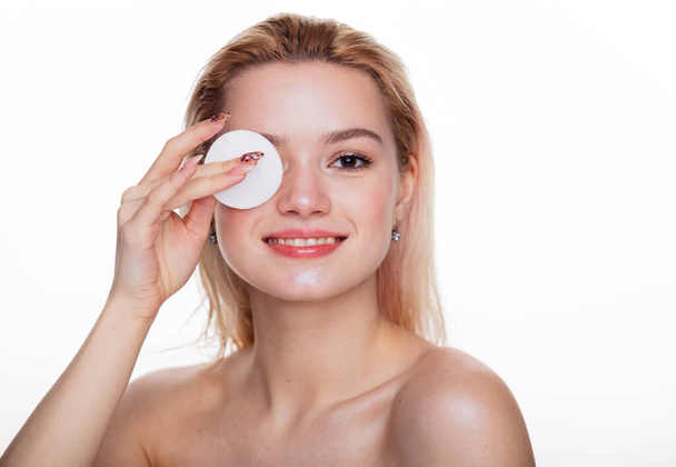 Cute beautiful natural blonde cleaning face with cotton sponge. Gorgeous woman face with nude make up and naked shoulders with cleaning sponge close to face over white background, skin care concept - Photo, Image