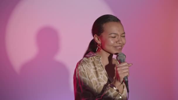 Średnie powolne ujęcie wesołej młodej birasowej kobiety śpiewającej w mikrofonie i poruszającej się energicznie na różowym tle - Materiał filmowy, wideo