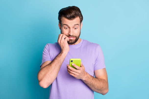Foto retrato del hombre asombrado manteniendo el teléfono inteligente mordiendo los dedos mirando el problema aislado vívido fondo de color azul - Foto, imagen