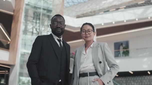 Tracking portrait shot of African-American businessman and Asian businesswoman in formal wear and glasses standing in office building lobby and smiling for camera - Filmagem, Vídeo