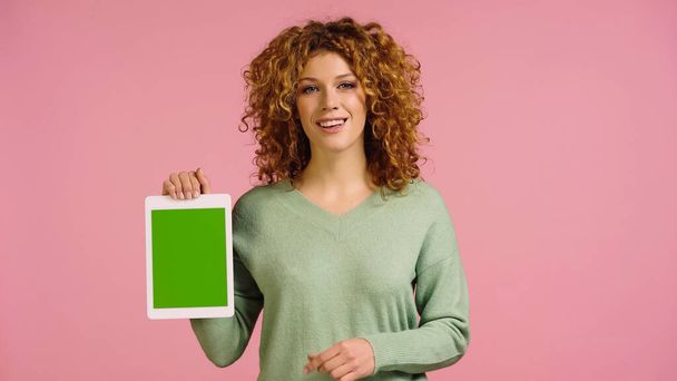 šťastná zrzka žena v zeleném jumper drží digitální tablet se zelenou obrazovkou izolované na růžové - Fotografie, Obrázek