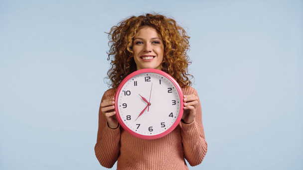 cheerful redhead woman holding white round clock while looking at camera isolated on blue - Photo, Image