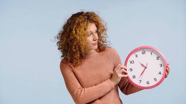 surprised redhead woman looking at round clock isolated on blue - Φωτογραφία, εικόνα