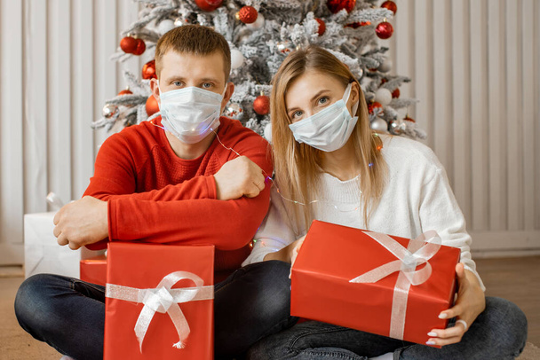 Couple man and woman with hygiene mask sit close to each other and look at camera for celebrating of Christmas during pandemic of Covid-19. - 写真・画像