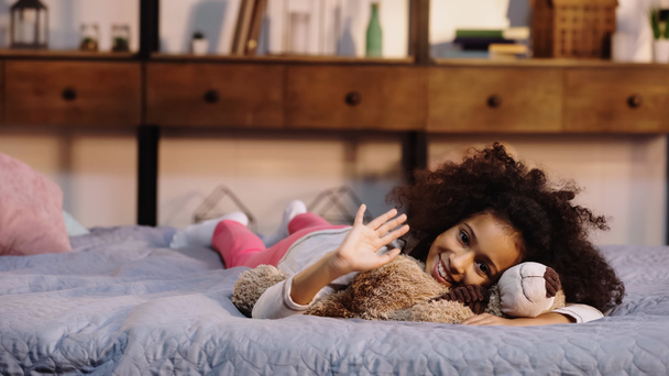 happy african american child waving hand while lying with teddy bear in bed - Fotoğraf, Görsel