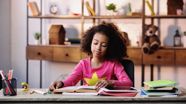 african american girl reading book near copybooks and digital tablet on desk - 写真・画像