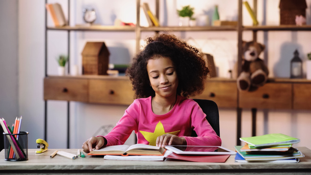 机の上のノートとデジタルタブレットの近くに幸せなアフリカ系アメリカ人の女の子の読書本 - 写真・画像