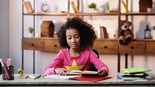 щаслива афроамериканська дівчина читає книгу біля блокнотів на столі
 - Фото, зображення