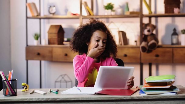 encaracolado menina americana africana usando tablet digital enquanto cobre a boca perto de cadernos na mesa  - Foto, Imagem
