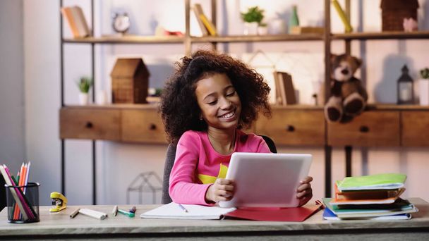 vrolijk Afrikaans amerikaans meisje met behulp van digitale tablet in de buurt notebooks op bureau  - Foto, afbeelding