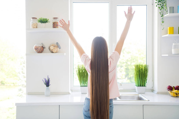 Πίσω πίσω όψη της ελκυστικής μακρυμάλλης κορίτσι αυξάνεται τα χέρια μέχρι ανάπαυσης στο σπίτι φως λευκή κουζίνα σε εσωτερικούς χώρους - Φωτογραφία, εικόνα