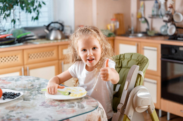 Happy little girl child show gesture of approval, Thumbs up by finger after eating food in kitchen at table sitting On baby food chair. Lunch of cheerful kid Caucasian blonde 5 years - Photo, image
