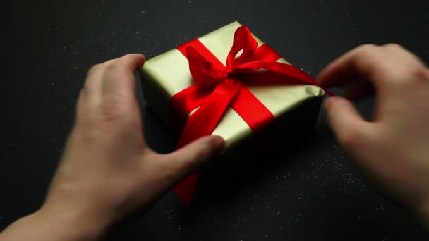 Ouverture d'un cadeau festif enveloppé dans de l'or et un arc rouge sur un fond noir. Un cadeau agréable et une surprise pour vos proches. - Séquence, vidéo