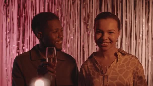 Tiro médio de feliz jovem casal afro-americano com taças de champanhe nas mãos fazendo brinde na câmera de pé contra cortina franja folha vermelha na festa de Ano Novo - Filmagem, Vídeo